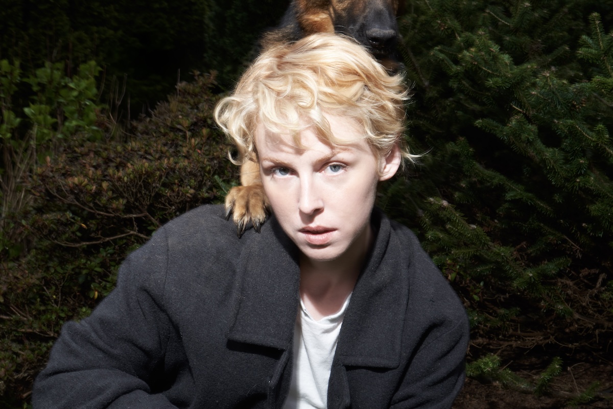 Portrait of singer-songwriter Fenne Lily in a black coat with a German Shepherd's paw resting on her shoulder outdoors.
